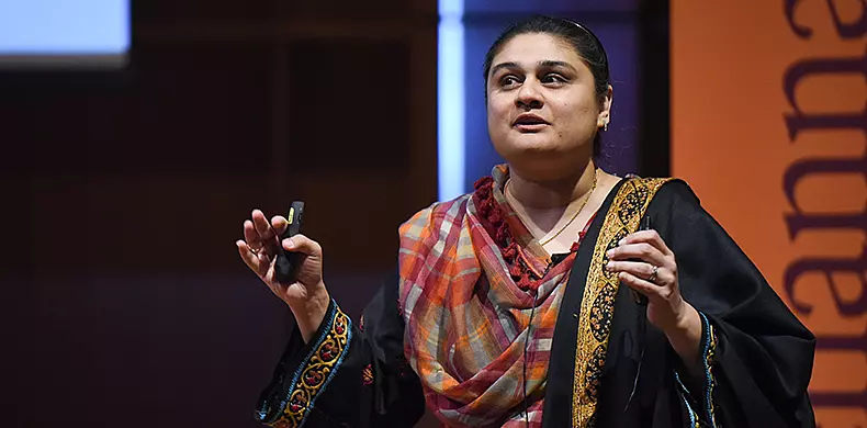 A woman in a colorful traditional shawl speaks on stage, holding a remote control in one hand and gesturing with the other, with a orange and black background behind her.