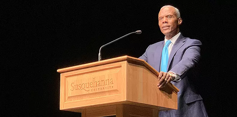 A man in a suit and blue tie stands at a wooden podium with 