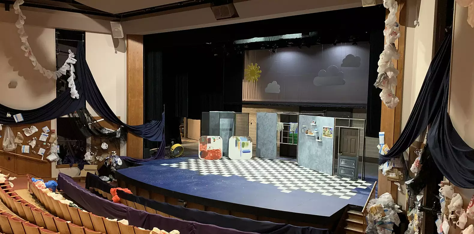 A theater stage set for a play features a checkered floor, several painted wall panels, and props resembling kitchen appliances. The auditorium has rows of empty seats and decorative elements hanging along the walls.