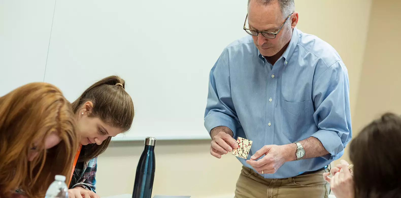 A man in glasses and a blue shirt stands at a table, holding a patterned card, while three women seated nearby focus on their work. A metal water bottle and papers are on the table in a brightly lit room.