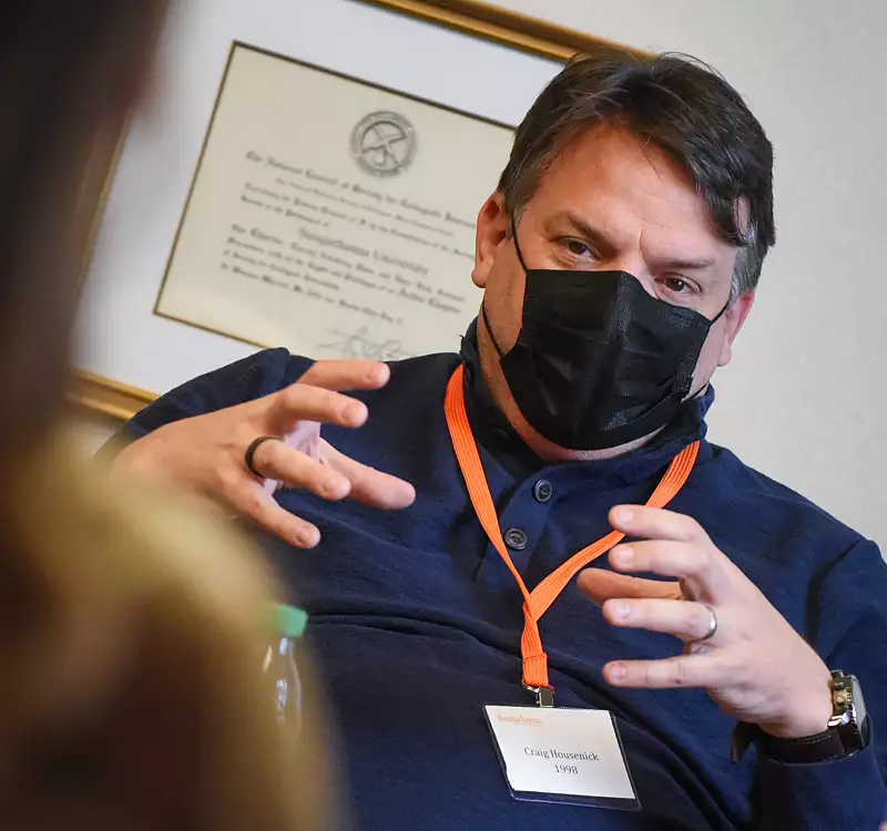 A man wearing a black mask and orange lanyard speaks, gesturing with his hands. A blurred person is in the foreground, and a framed certificate hangs on the wall behind him.