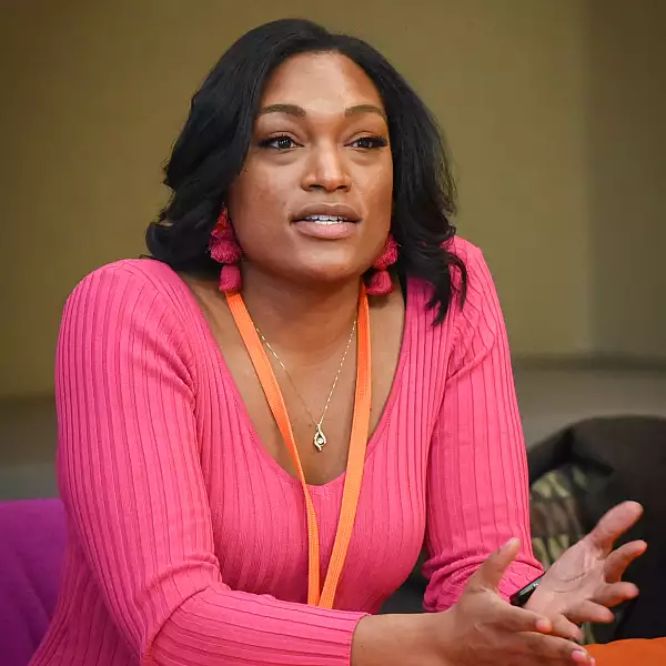 A woman with long dark hair, wearing a bright pink sweater and matching tassel earrings, gestures with her hands while speaking. She has an orange lanyard around her neck and is sitting indoors.