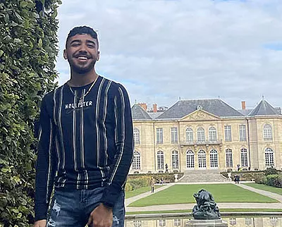 A man in a striped Hollister shirt stands smiling in front of a large historic building with manicured gardens and a sculptural fountain. The sky is partly cloudy.