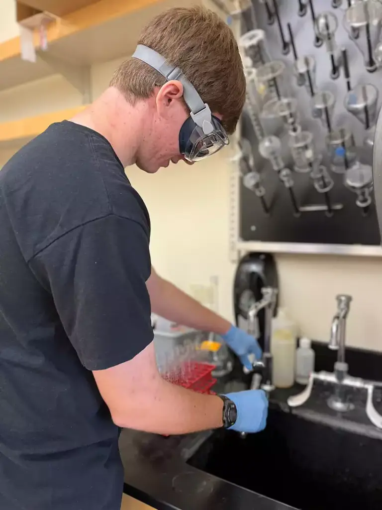 A person wearing safety goggles and gloves is working at a laboratory sink. They are rinsing test tubes under a faucet. A rack with inverted glass containers is visible in the background.