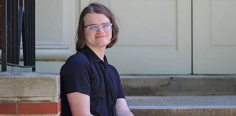 Person with shoulder-length hair and glasses, wearing a black shirt, seated on outdoor steps near a building with a light-colored door, smiling at the camera.