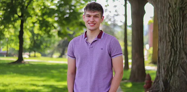 A young man in a purple polo shirt smiles while standing outdoors. The background features lush green trees and grass, suggesting a park or garden setting.