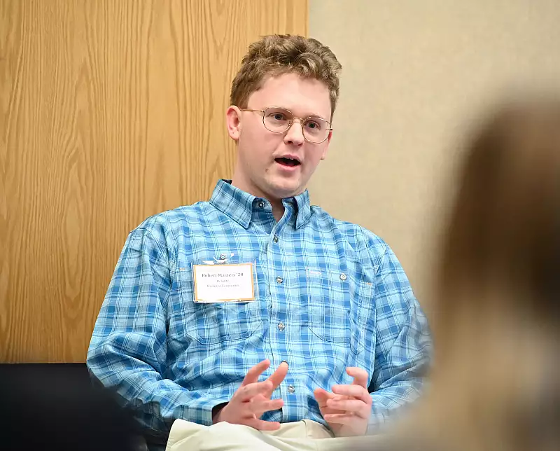 A person wearing glasses and a blue plaid shirt speaks while gesturing with their hands. They are seated against a light-colored wall with a wooden panel behind them, and a name tag is visible.