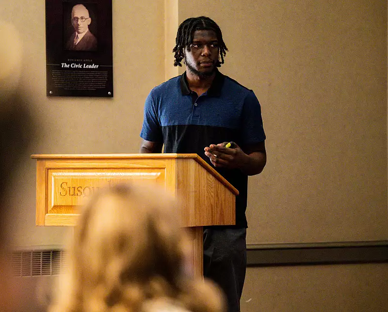 A person with dreadlocks, wearing a blue and black polo shirt, stands behind a wooden podium labeled 