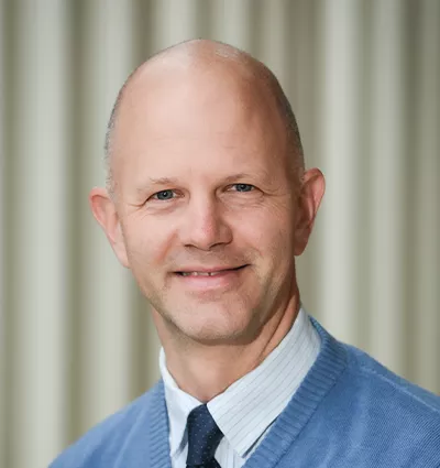 A smiling person with a bald head is wearing a blue sweater over a collared shirt and tie. The background features vertical, beige stripes.
