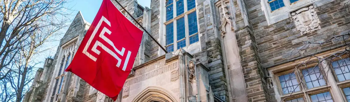 A red flag with a white emblem hangs outside a stone building with large windows and intricate architectural details. Bare tree branches are visible to the left.