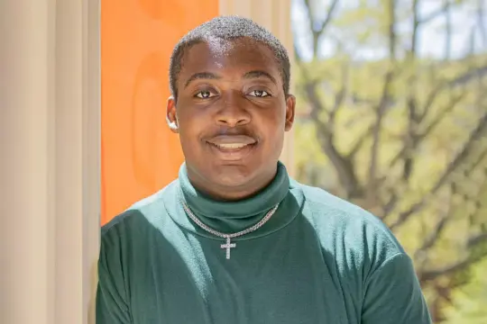 A computer science student wearing a green turtleneck and a cross necklace, standing indoors by a window with a view of trees outside.