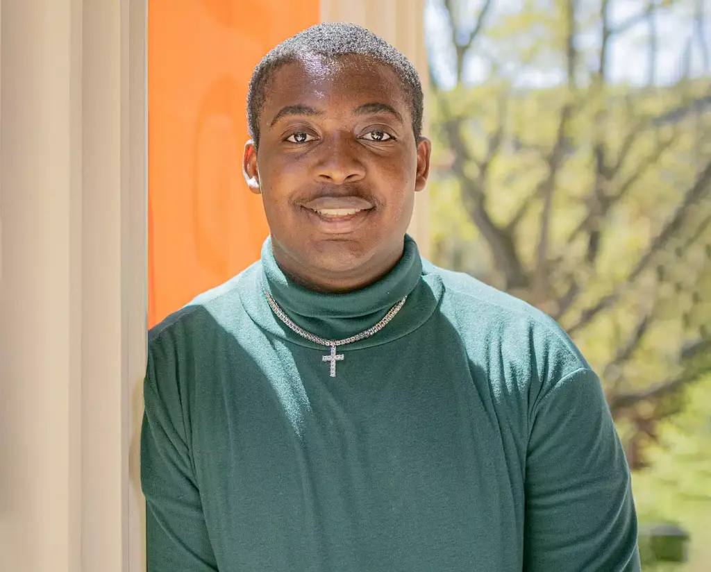 A person stands smiling next to an orange wall, wearing a green turtleneck and a cross pendant necklace. Sunlight and blurred trees are visible in the background.