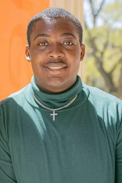 A person stands smiling next to an orange wall, wearing a green turtleneck and a cross pendant necklace. Sunlight and blurred trees are visible in the background.