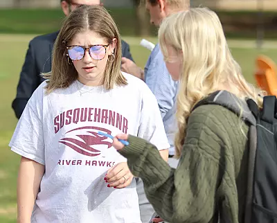 Two people are outdoors. One wears a 体育买球 River Hawks T-shirt and sunglasses, looking at something. The other has long hair and a green sweater, holding a pen and paper. Blurred people in the background suggest a campus or park setting.