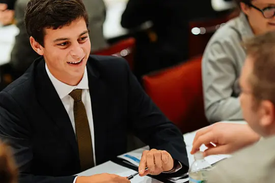 A young man in a suit smiles while seated at a table, engaged in conversation with others. Papers and a water bottle are visible on the table. The setting appears to be a professional or formal event.