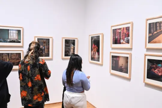 People are standing in a gallery, observing framed photographs on white walls. The artworks feature various scenes and subjects. The group includes individuals engaged in viewing and discussing the photographs.