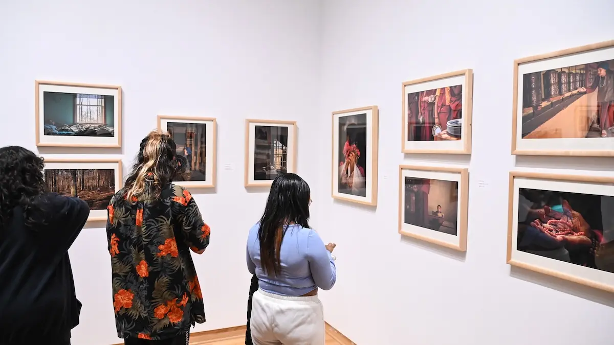 People are standing in a gallery, observing framed photographs on white walls. The artworks feature various scenes and subjects. The group includes individuals engaged in viewing and discussing the photographs.