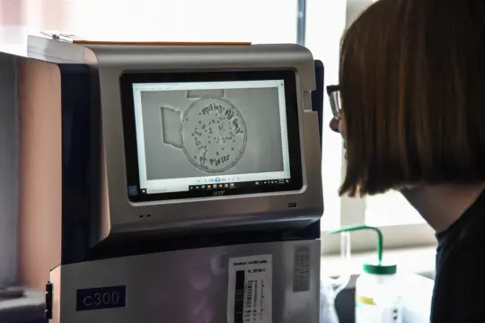 A person with shoulder-length hair and glasses examines a computer screen displaying a lab image, possibly a petri dish. The scene appears to be in a laboratory setting with various equipment around.