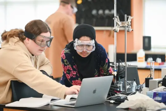 Two students wearing safety goggles work together at a lab table with a laptop and scientific equipment. One student wears a hijab and floral dress. The lab is bright, with various tools and papers on the table.