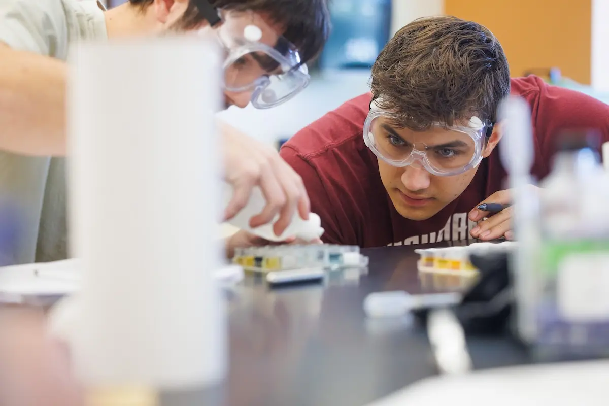 Two students wearing safety goggles work intently on a science experiment at a chemistry lab table. One student carefully drops liquid into a container, while the other observes.