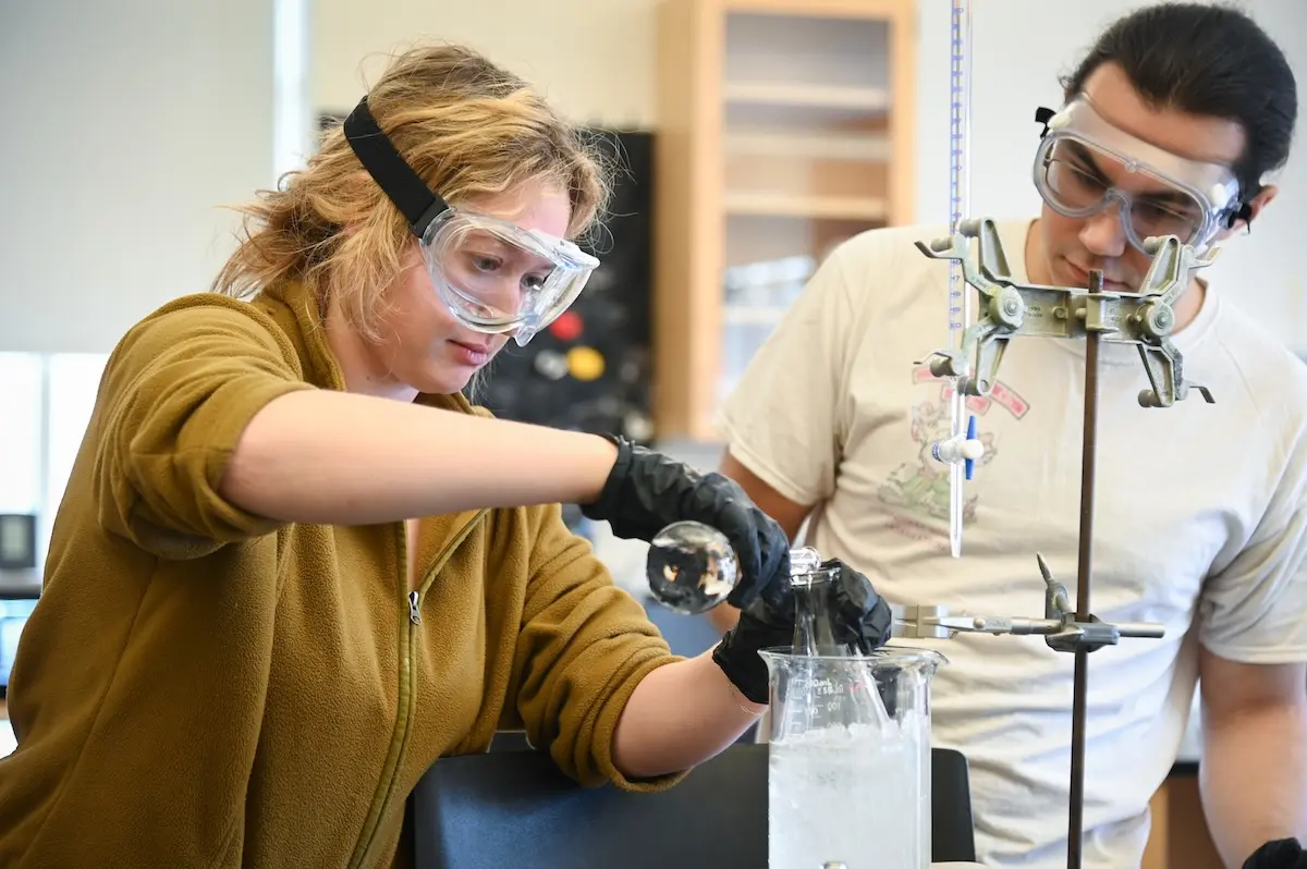 Two individuals in a laboratory setting conduct an experiment. One person carefully pours a liquid from a flask into a beaker, while the other observes. Both wear protective goggles and gloves, and a burette is clamped to a stand nearby.