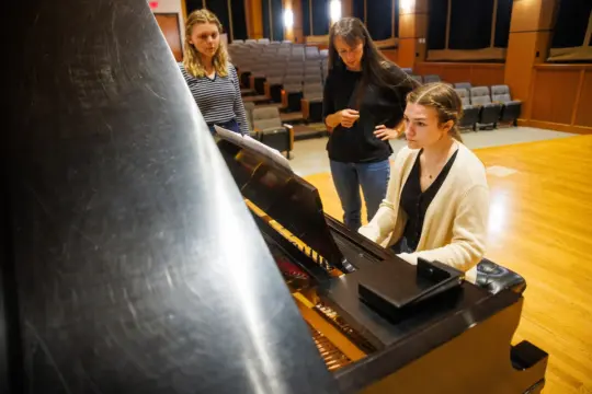 Three women in a music hall; one sits at a grand piano playing, while the other two stand nearby, listening attentively. The hall is empty, with rows of seats visible in the background.