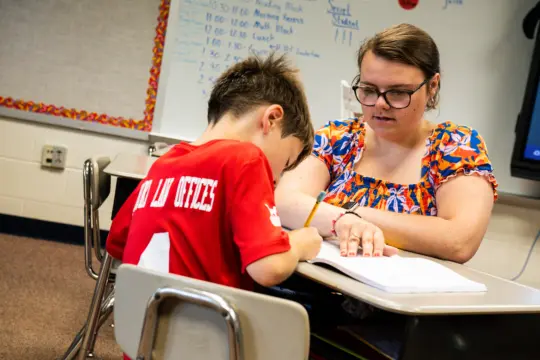 A teacher with glasses and a colorful blouse is sitting at a desk, assisting a young boy in a red shirt with writing. The classroom has a whiteboard with a schedule written on it in the background.