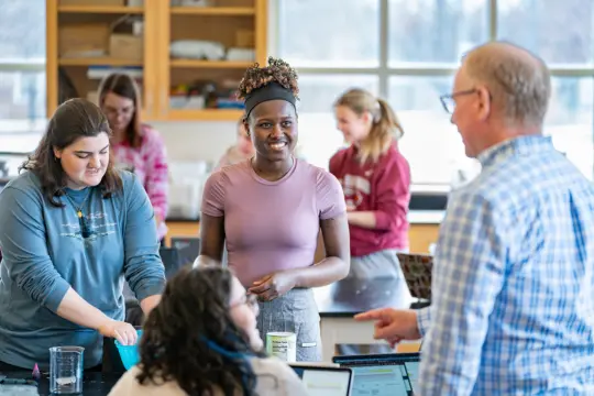 A classroom scene with a teacher and students engaging in a science activity. Students are gathered around a table with various materials, while the teacher, in a blue checked shirt, is speaking to them. Shelves in the background hold lab equipment.