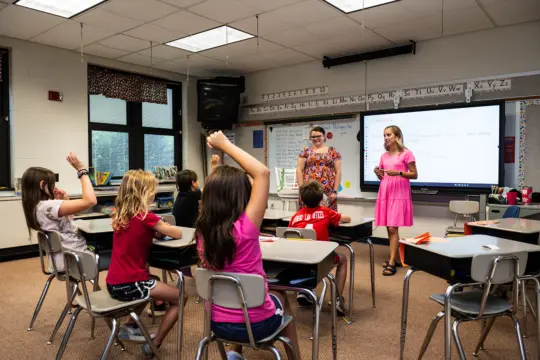A classroom with children seated at desks. One child raises their hand. Two adults stand at the front; one is teaching, the other is observing. A whiteboard displays text. The room is bright with natural light from large windows.