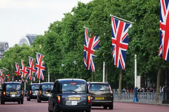 A row of black taxis drives down a wide street lined with large Union Jack flags on tall poles. The street is bordered by lush green trees.