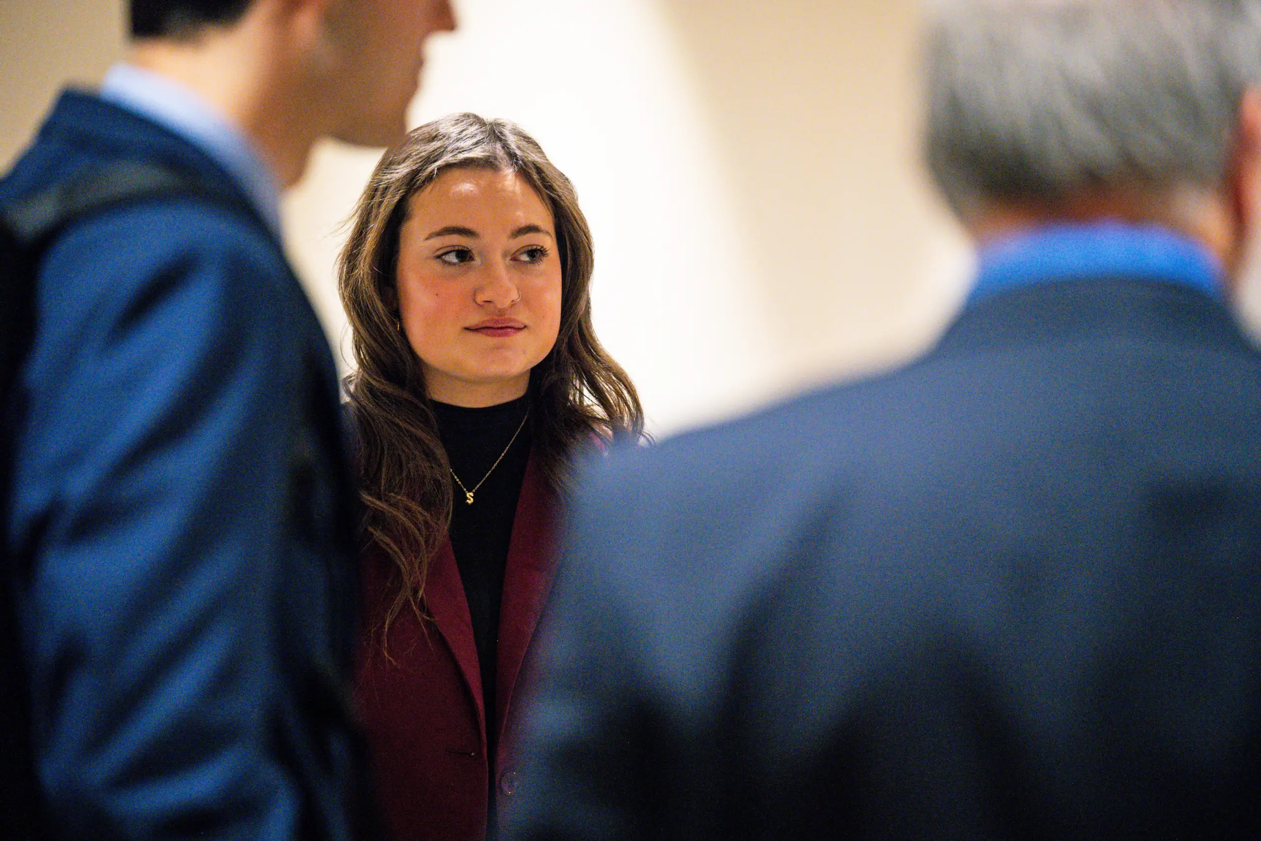 A woman with long hair wearing a maroon blazer stands among a group of people. She is looking slightly to the side with a thoughtful expression, perhaps contemplating the next step in her MBA 4+1 Program journey. The image is indoors with a neutral background.