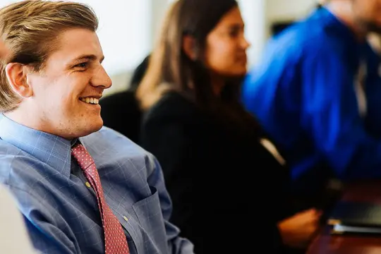 A group of people sitting at a table in a meeting room. A man in the foreground, wearing a blue shirt and pink tie, is smiling and appears engaged. Others, slightly blurred, sit beside him in professional attire.