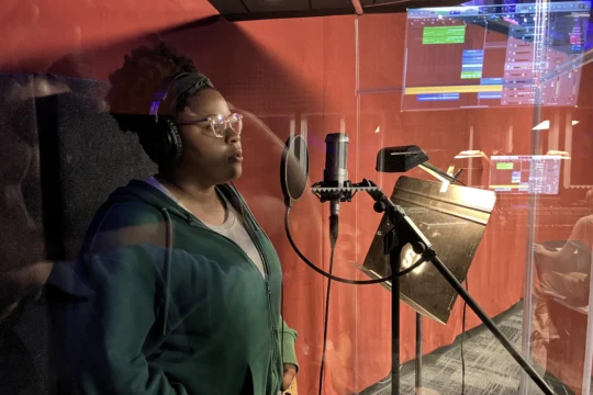 A person wearing headphones and a green hoodie stands in a recording booth, singing or speaking into a microphone. Music production software glows on monitors through the glass window, highlighting the creative process.