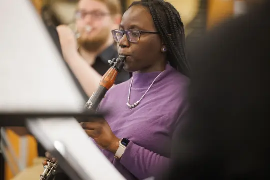A woman wearing a purple sweater plays a woodwind instrument, focusing intently. She has braided hair and wears glasses and a necklace. A blurred figure is playing an instrument in the background.