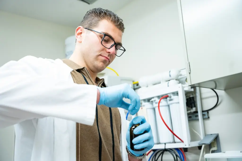 A person in a lab coat and gloves is handling a small container in a laboratory setting, focused on their task. Equipment is visible in the background.