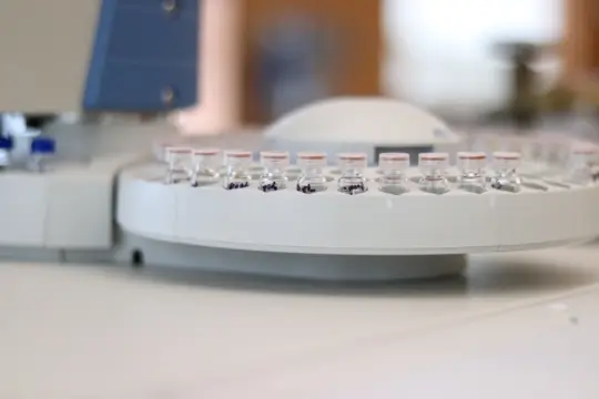 A close-up of several small glass vials with labels in a circular holder. The container is part of a laboratory instrument, likely for chemical or medical analysis. The background is blurred, emphasizing the vials.