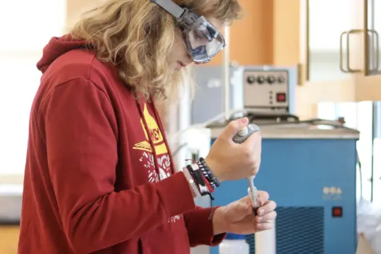 A person with long hair is wearing a red hoodie and safety goggles, using a pipette in a laboratory. The background shows lab equipment and an orange wall.