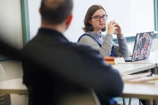 A person wearing glasses sits at a desk with an open laptop covered in stickers. They are looking attentively at someone blurred in the foreground. There are books on the desk, and a window in the background.