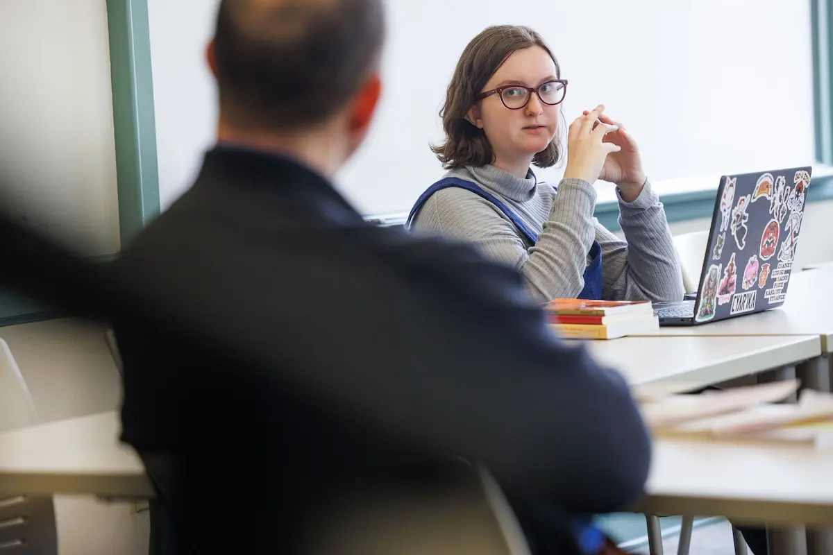 A person wearing glasses sits at a desk with an open laptop covered in stickers. They are looking attentively at someone blurred in the foreground. There are books on the desk, and a window in the background.