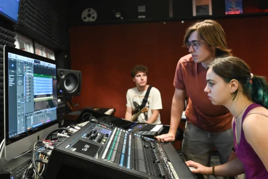 Three young people work in a music studio, immersed in music technology. Two focus intently on a mixing console and computer screen with audio tracks, while the third sits nearby with a keyboard. The room features soundproofing and audio equipment.