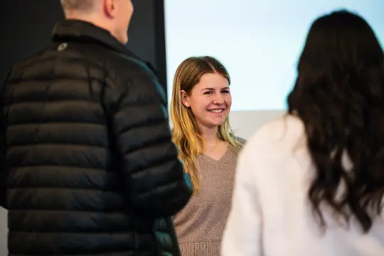 Three people are standing and conversing in a room. A woman with long blonde hair is smiling while engaging with two others, one wearing a black jacket and the other with long dark hair. The background hints at a blurred white and black screen, reminiscent of a secondary education seminar setting.