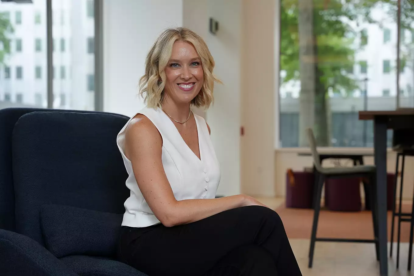 A woman with blonde, wavy hair wearing a white sleeveless blouse and black pants sits smiling on a dark chair in a modern, bright office space—an ideal setting for Advertising and Public Relations professionals—with large windows and greenery outside.