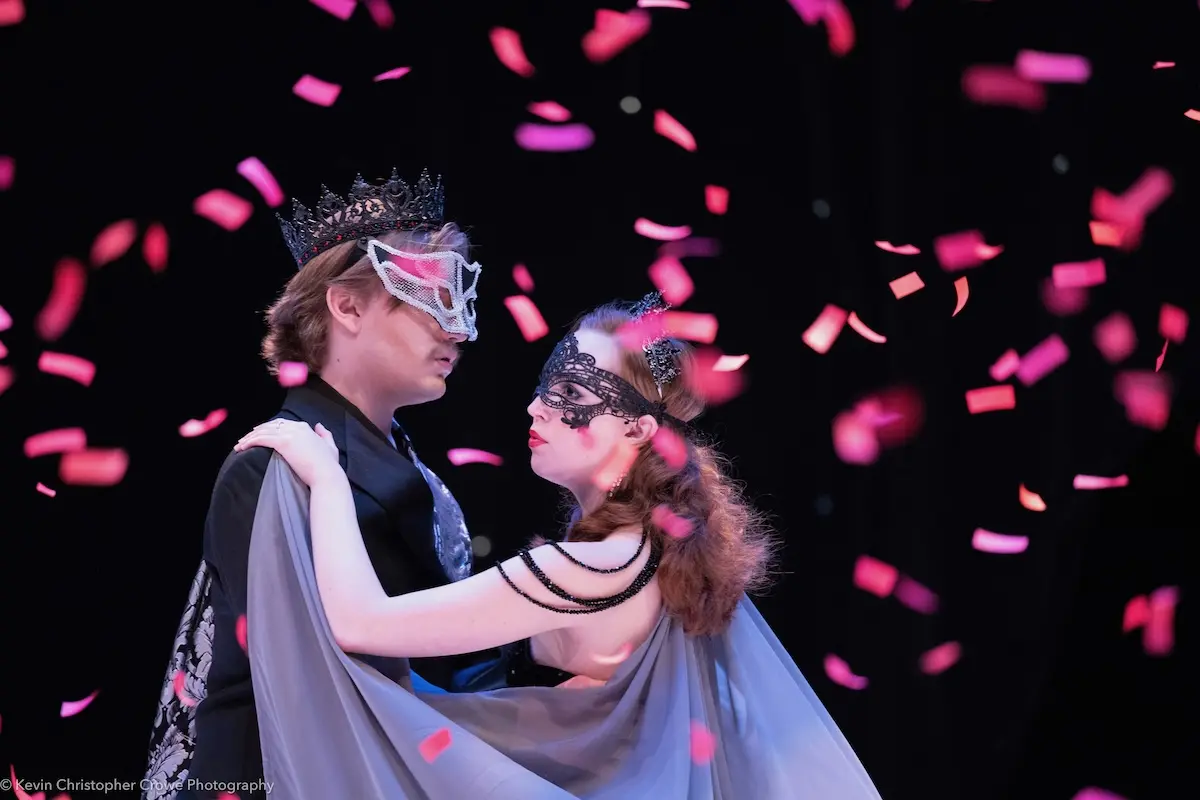 A couple wearing elegant masquerade masks and formal attire dances under a shower of pink confetti. They are on a stage, with the man wearing a crown and the woman draped in a flowing cape. The scene is set against a dark background.