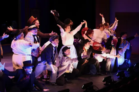 A theater group dressed in colorful, Victorian-era costumes performs energetically on stage. They strike dynamic poses with arms outstretched, engaging with the audience under dramatic lighting.