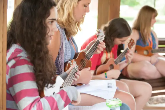 Four girls sit closely together outdoors, playing ukuleles and reading from sheet music, embodying the joy of music education. The focus is on the girl in the foreground with curly hair and a striped shirt; the background is softly blurred.