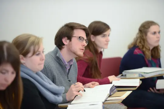 A group of students sits attentively in a classroom, taking notes on applied linguistics. They hold pens and notebooks, focusing intently on the lesson. The background is a plain white wall, creating a simple, academic setting perfect for exploring this intricate field of study.