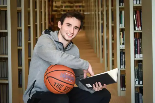 A young man in a gray hoodie kneels in a library aisle, smiling at the camera. He holds a black book in one hand and a basketball in the other, surrounded by shelves of books.