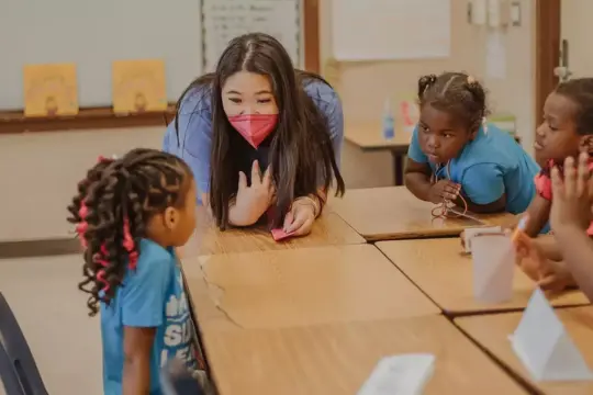 A teacher in a pink mask connects with four young students seated at a wooden table, embodying the principles of Studies in Early Education. The children, dressed casually, listen attentively amidst classroom decorations and a whiteboard backdrop.