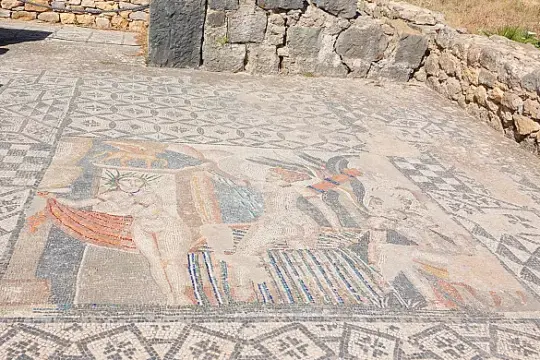 Ancient mosaic depicting three figures, surrounded by intricate geometric patterns. The scene includes vibrant colors and is set against a background of stone walls and grass.