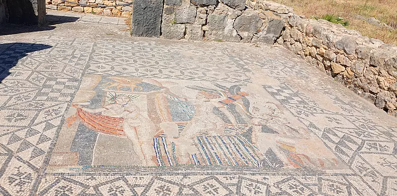 Ancient mosaic depicting three figures, surrounded by intricate geometric patterns. The scene includes vibrant colors and is set against a background of stone walls and grass.